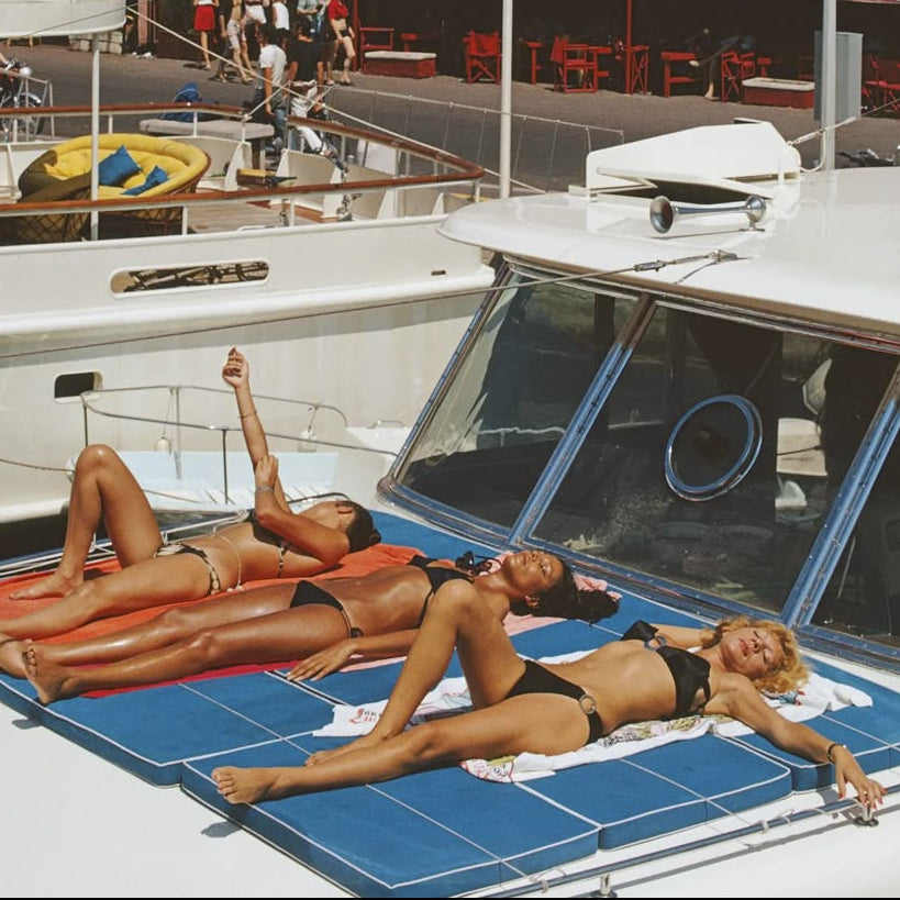 Women sunbathing in black bikinis on a yacht in Saint-Tropez harbour, with colourful waterfront buildings and café terraces in the background