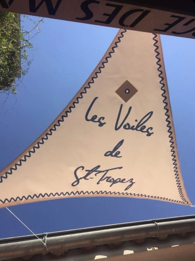 Triangular sail shade reading "Les Voiles de St-Tropez" against a blue sky, at Shellona beach club in Saint-Tropez