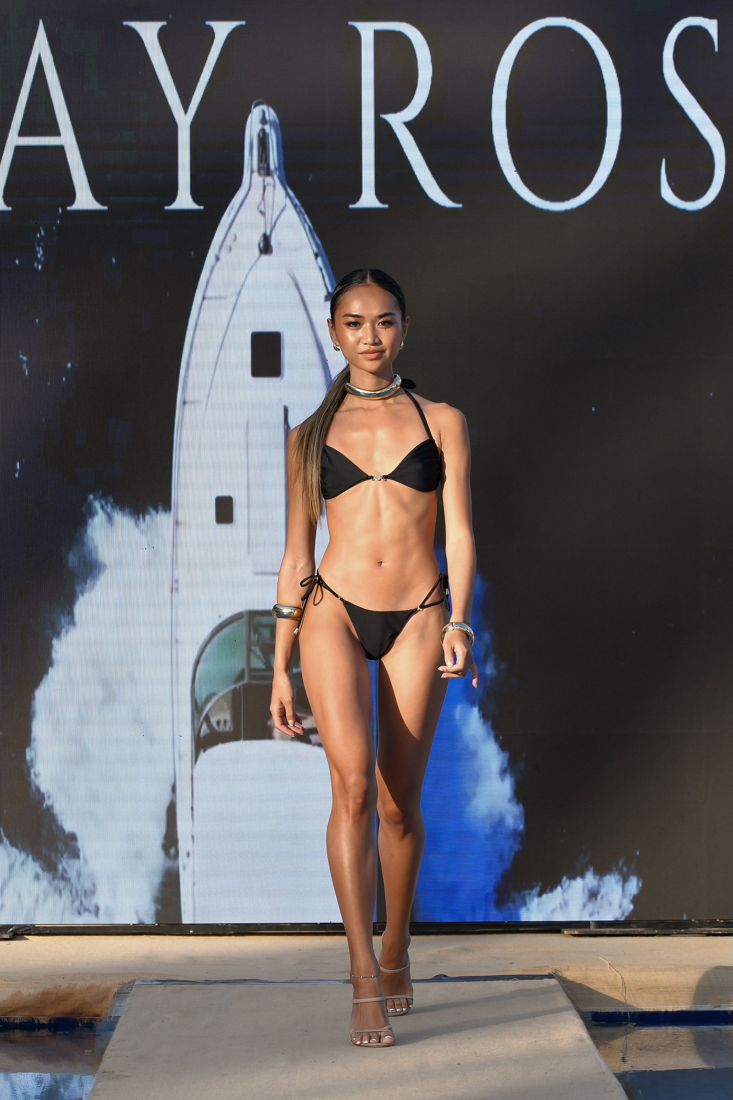 Model walking at Ibiza Swim Week 2025 wearing the Chia bikini set in ebony black by May Rose Swimwear, featuring a string bottom with adjustable double straps and gold ring detail, and a bikini top with central 18k gold-plated charm